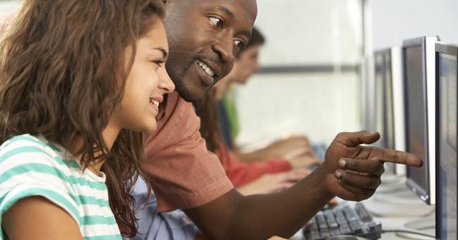 Teacher Helping Students Working At Computers In Classroom
